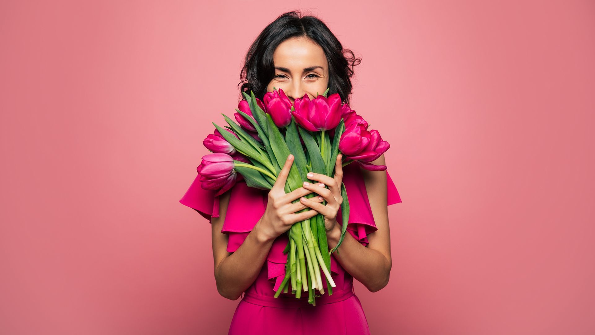 International Women's Day. Extremely happy woman in a bright pink dress is smelling a bunch of spring flowers, which she is holding in her hands.